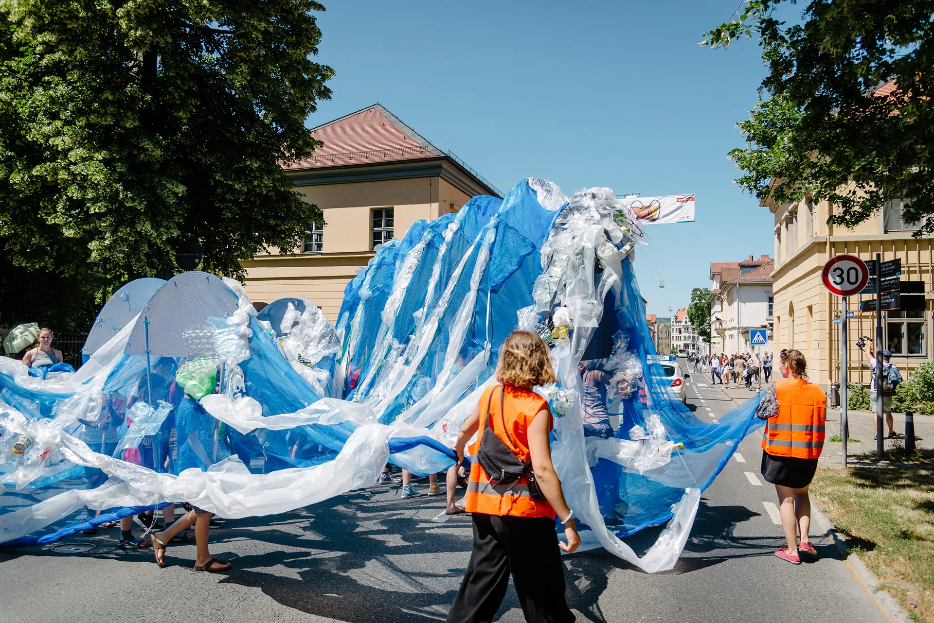 Die Welle aus Plastikfolie in Bewegung auf einer sonnigen Straße, begleitet von Ordner*innen