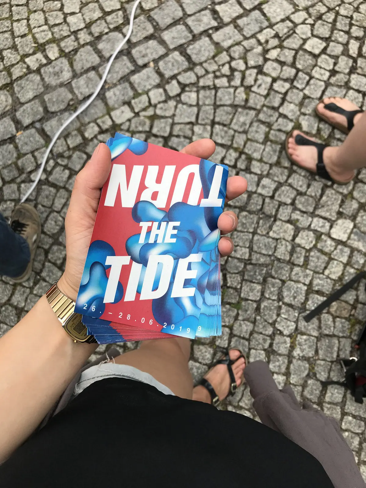 Hand hält einen Flyer mit dem Titel „Turn the Tide“ über Kopfsteinpflaster