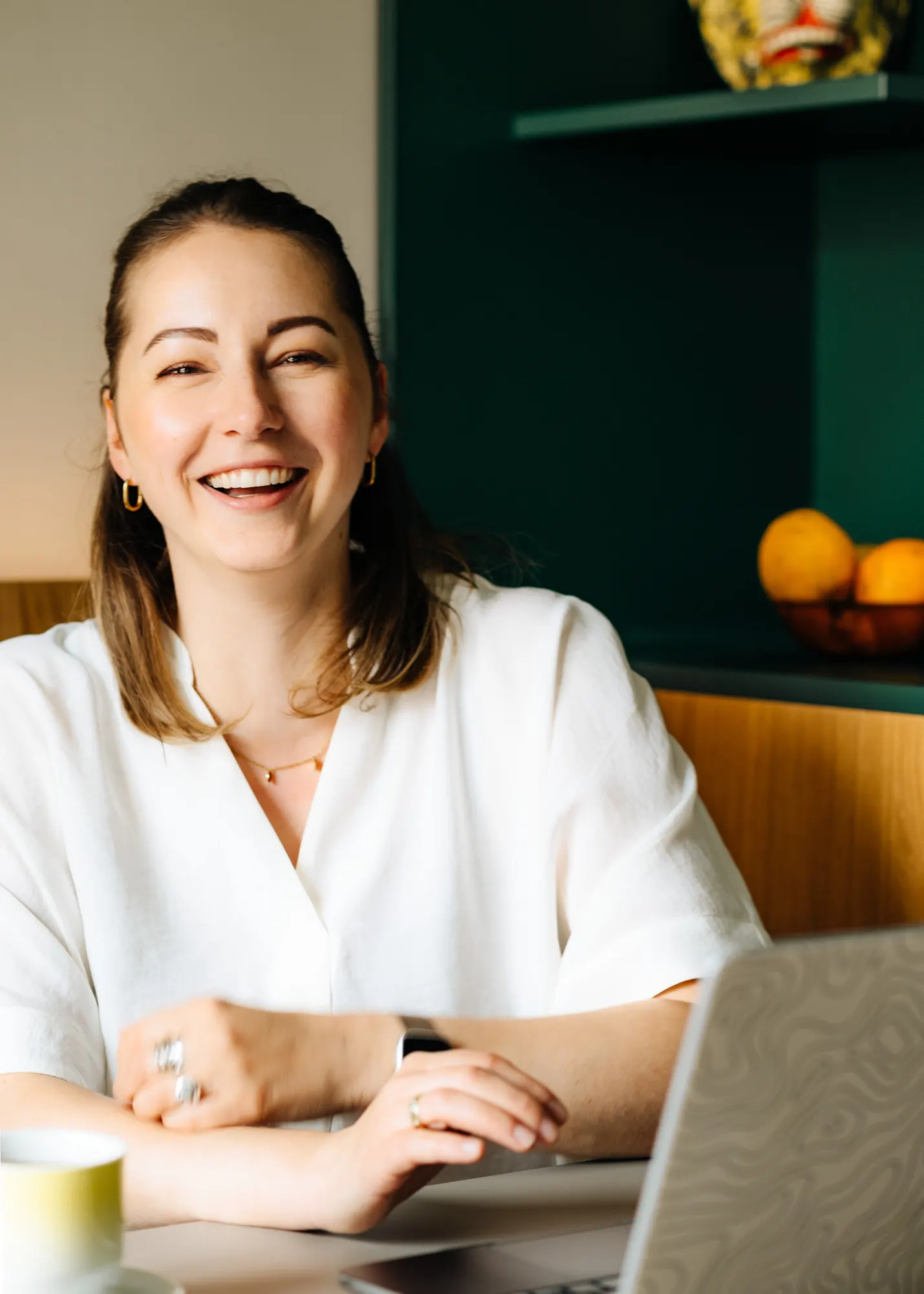 Anne Lutterberg sitzt lachend an einem Tisch mit Laptop und Tasse, im Hintergrund ein dunkelgrünes Regal
